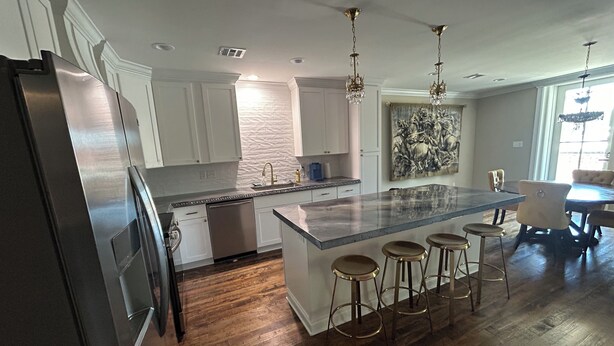 A modern kitchen with white cabinets, a stainless steel refrigerator, a marble island with bar stools, and a dining area with a large table and chairs.