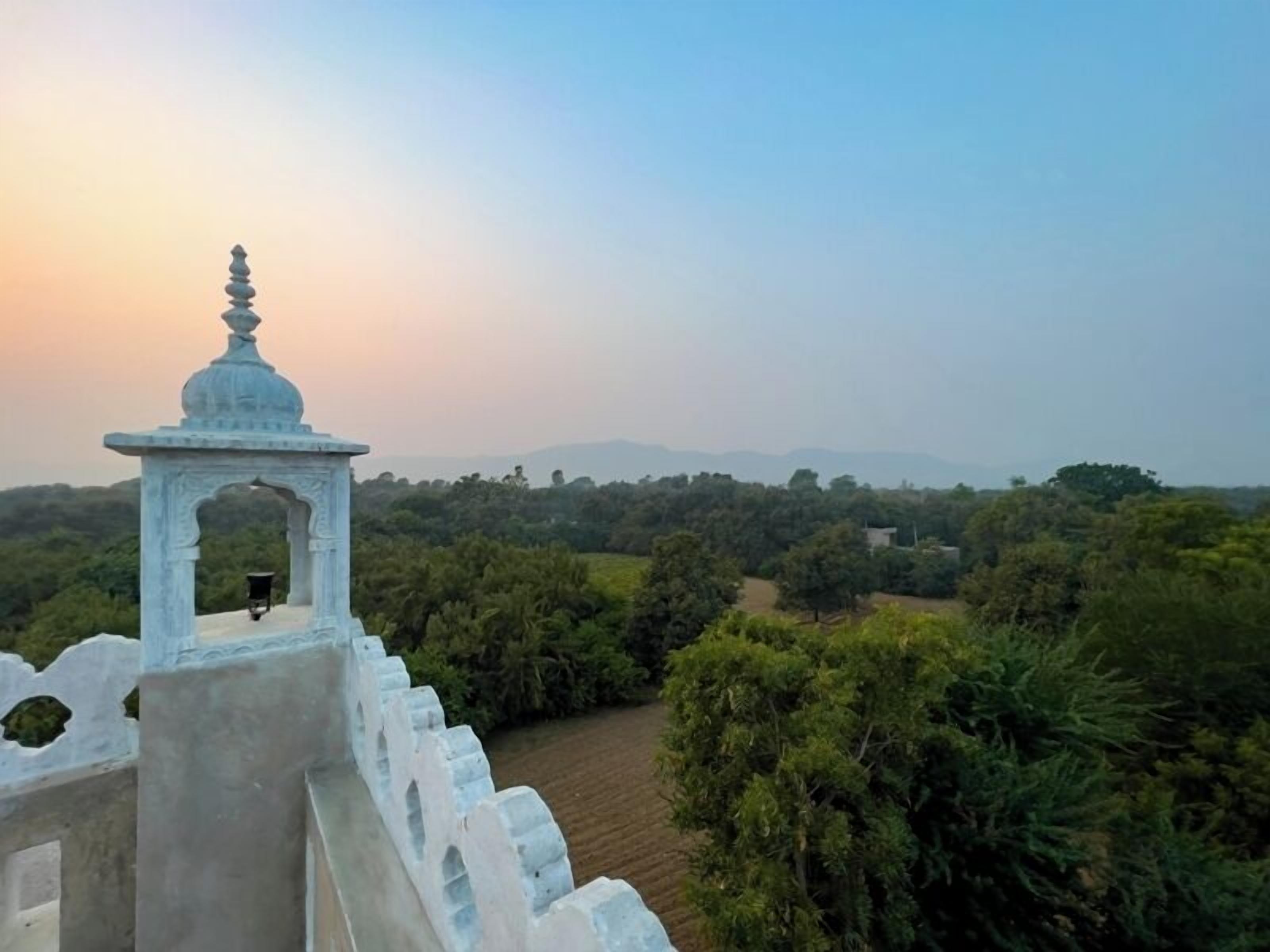 A white stone structure with a dome and archway, overlooking a lush green landscape with trees and distant hills.