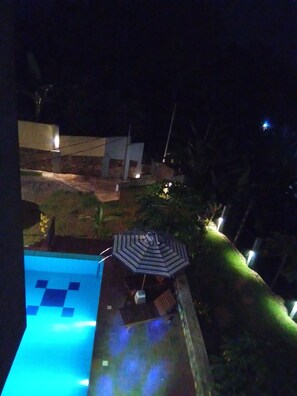A nighttime view of a pool area with a lounge chair and umbrella, surrounded by greenery and buildings. - The Moose Weligama (Weligama)