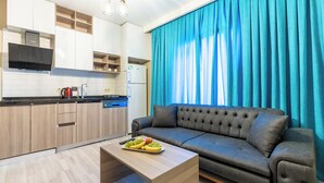 A modern kitchen with wooden cabinets, a built-in oven, and a coffee maker. A black leather sofa is positioned near a large window with blue curtains. - MELISA SUIT RESIDENCE (Istanbul)