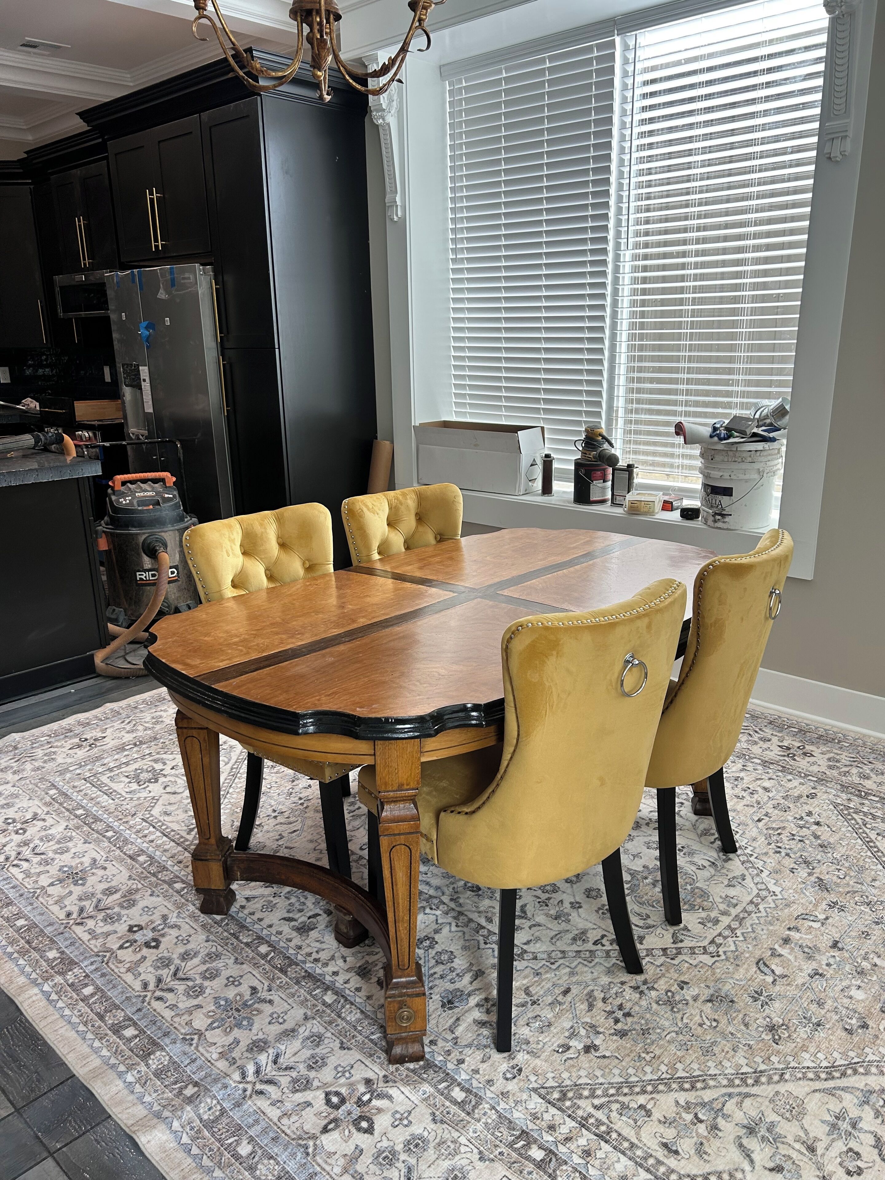 A dining area with a wooden table and yellow upholstered chairs, a patterned rug, and a kitchen in the background.
