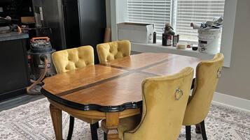A dining area with a wooden table and yellow upholstered chairs, a patterned rug, and a kitchen in the background.