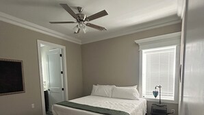 Un dormitorio con cama, ventilador de techo, ventana con persianas y mesita de noche.