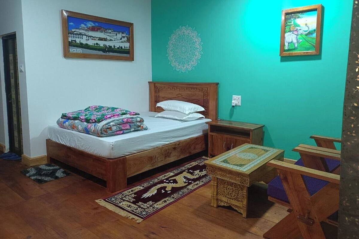 A bedroom with a wooden bed, a nightstand, a small table, and a chair. There is a picture on the wall and a rug on the floor.