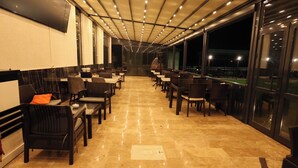 A spacious indoor area with rows of chairs and tables, large windows, and a ceiling with linear lights.