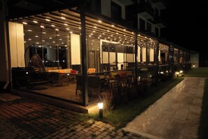 Een terras met tafels en stoelen, een overdekt zitgedeelte en een glazen constructie met verlichting.