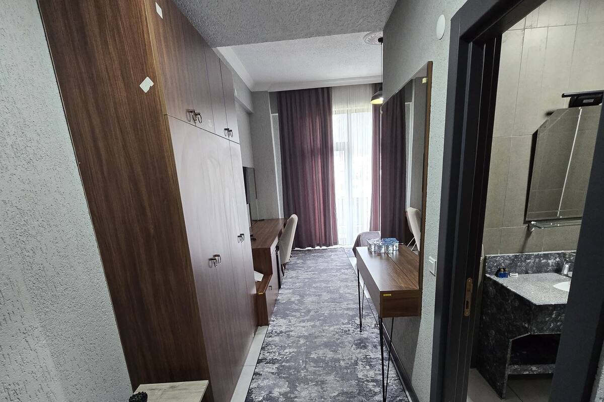 Een hotelgang met een houten garderobe, een klein tafeltje met een stoel, een bureau met een stoel en een badkamer met een wastafel en spiegel.