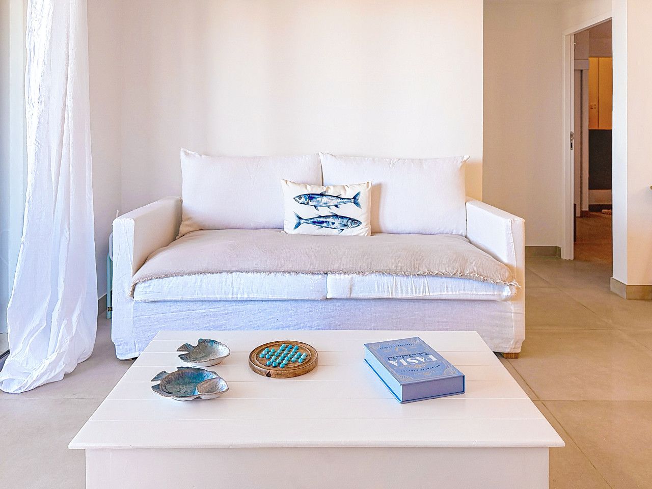 Un canapé blanc avec deux coussins, une table basse blanche agrémentée d’objets décoratifs et une porte donnant sur une autre pièce.