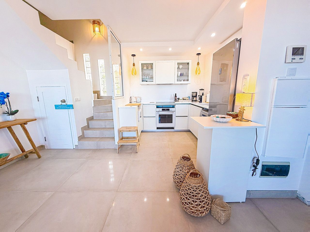 A modern kitchen with white cabinets, a built-in oven, and a refrigerator. There is a wooden stool and a woven basket on the floor.