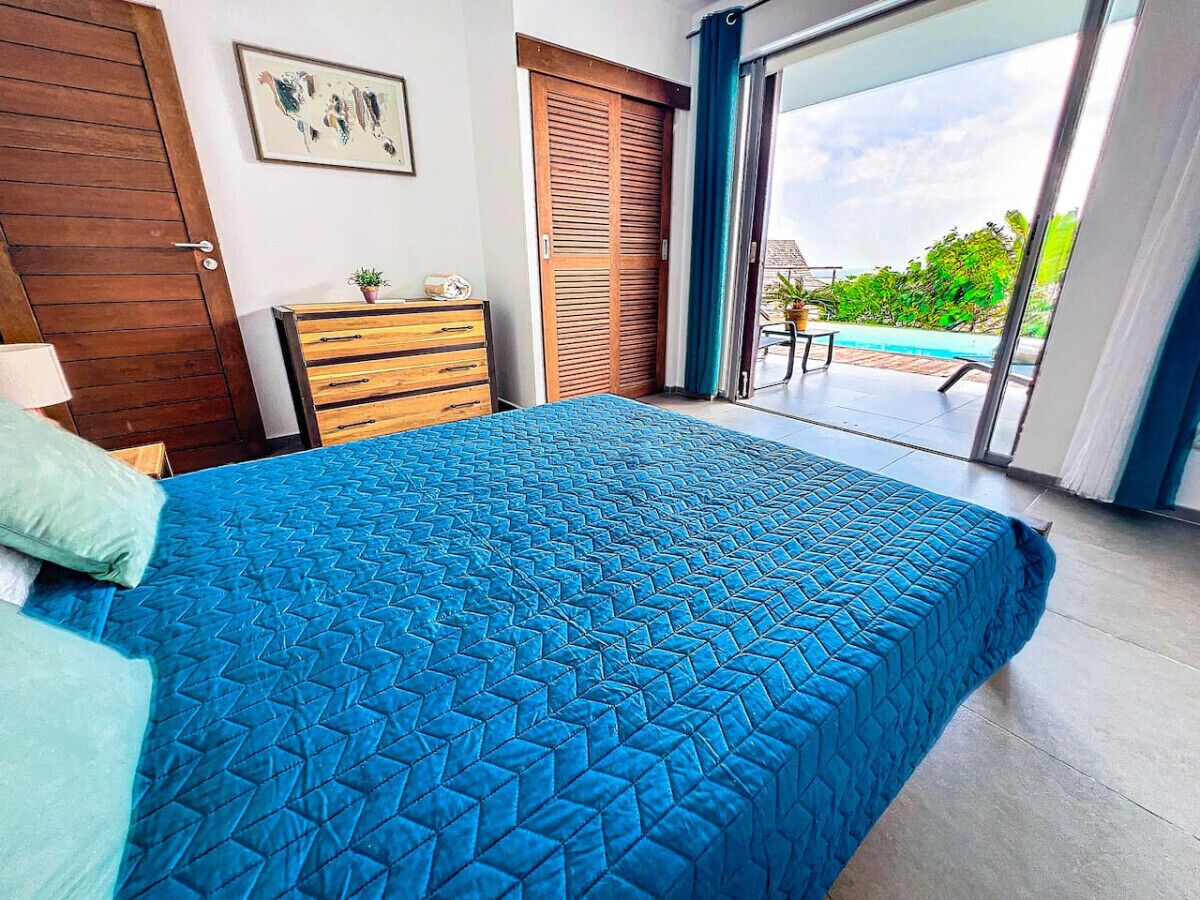 A bedroom with a large bed, a wooden dresser, a door, and a view of a pool and greenery.