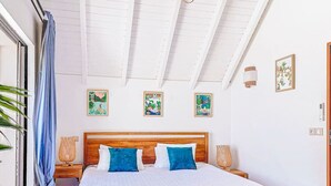Ein Schlafzimmer mit einem hölzernen Bett, weißer Bettwäsche und blauen Kissen. Zwei Wandlampen und gerahmte Bilder sind an der Wand angebracht.