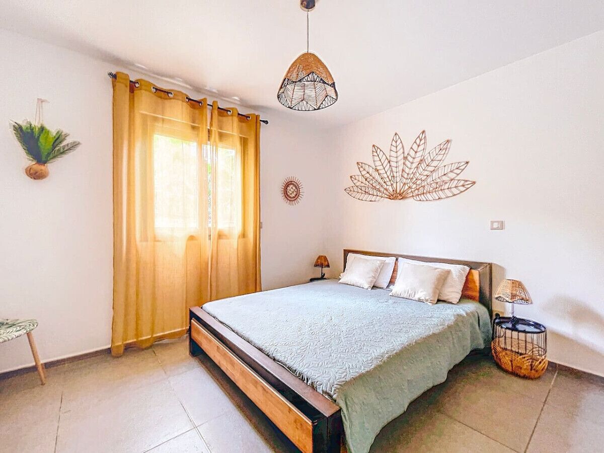 Une chambre à coucher avec un lit, des rideaux, un élément de décoration murale, une lampe de chevet et une petite table sur laquelle est posée une plante.