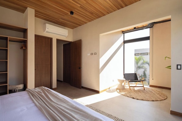 A modern bedroom with a large bed, a wardrobe, a chair by a sliding glass door, and an air conditioning unit.
