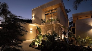 Una casa moderna de dos pisos con balcón, rodeada de plantas tropicales y un sendero que lleva a la entrada.