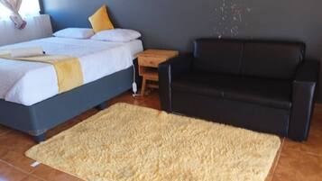 Un dormitorio con una cama, un sofá negro, una mesita de noche de madera y un tapete beige grande.