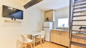 Cocina compacta con una televisión montada sobre una pequeña mesa y sillas, armarios de madera, refrigerador y una ventana con una planta en la encimera.