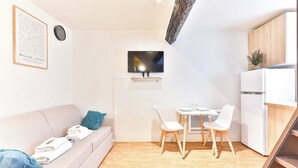 Un espacio habitable moderno y minimalista con sofá, mesa de comedor con sillas, televisor de pantalla plana en la pared, pequeña cocina con refrigerador y una escalera.