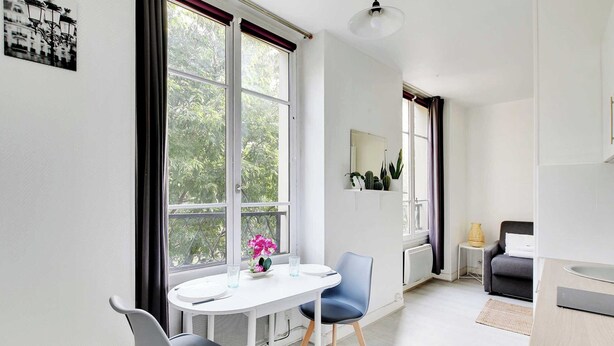 Une pièce moderne et bien éclairée, comprenant une table à manger, deux chaises, une petite table d’appoint, un canapé et une grande fenêtre donnant sur un paysage verdoyant.
