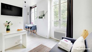 Une chambre d’hôtel moderne équipée d’une télévision à écran plat, d’un coin repas avec une table et des chaises, d’un canapé avec un porte-revues et d’une fenêtre avec des rideaux.