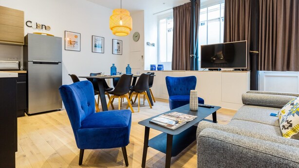 Ein modernes Wohnzimmer mit Sofa, Couchtisch und Fernseher. Eine Essecke mit Tisch und Stühlen ist ebenfalls vorhanden. Eine Küche inklusive Kühlschrank und Herd ist zu sehen.