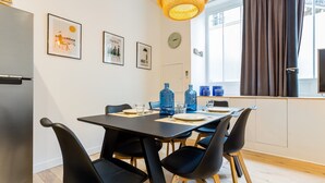 Ein moderner Essbereich mit einem schwarzen Tisch, vier schwarzen Stühlen und blauen Wasserflaschen. Links befindet sich ein Kühlschrank, ein Fenster mit Vorhängen sowie gerahmte Bilder an der Wand.
