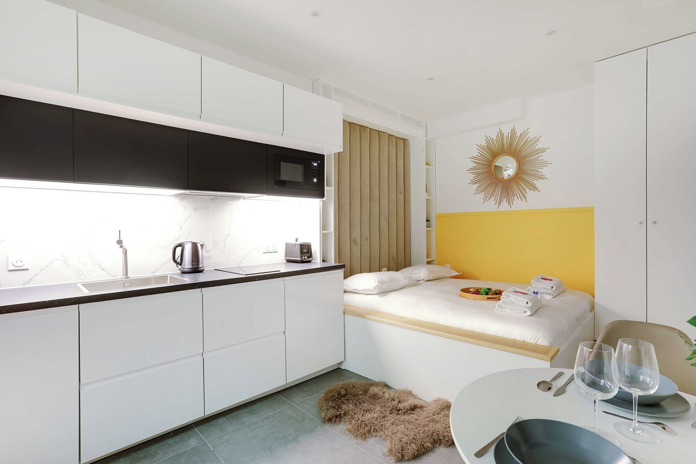 A compact hotel room with a kitchenette, a bed with a yellow headboard, a dining area with a round table, and a decorative sunburst mirror.
