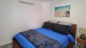 A bedroom with a bed, a wall-mounted air conditioner, a painting of a beach, and a small table with a lamp. - away on holiday (Terrigal)