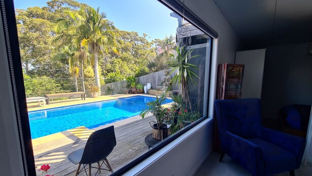 A room with a large window overlooking a swimming pool, a blue chair, and a wooden deck. - away on holiday (Terrigal)