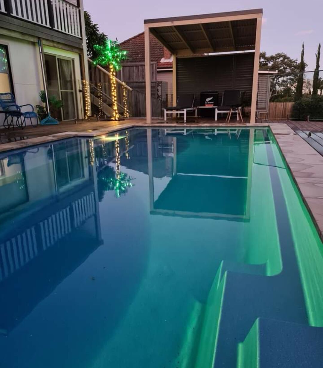 A swimming pool with clear water, surrounded by a covered patio area with chairs and a small table.