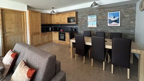 A modern kitchen with wooden cabinets, a dining area with a table and chairs, and a stone wall with a Val Morel poster. - Valmogrange E4 - 8 Persons - Luxury Apartment on the Piste (Les Avanchers-Valmorel)