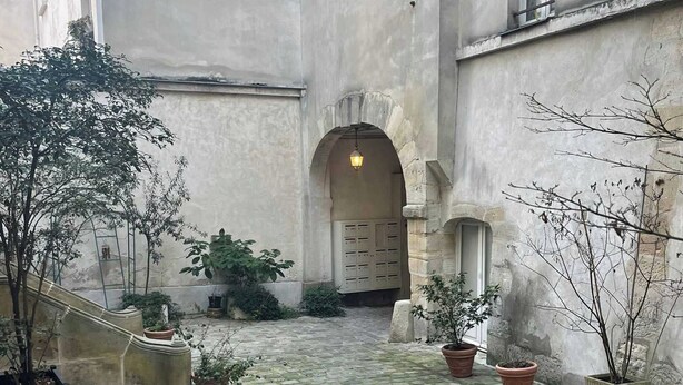 A courtyard with cobblestone flooring, potted plants, and a central archway leading to a building entrance. - Charming Duplex in Marais for 2 People (Paris)