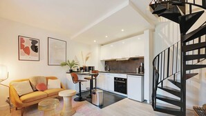A modern kitchen with white cabinets, a black spiral staircase, and a dining area with a yellow sofa and a pink rug. - Charming Duplex in Marais for 2 People (Paris)