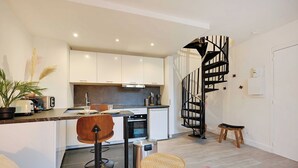 A modern kitchen with white cabinets, a black spiral staircase, a dining area with a wooden chair, and a small stool. - Charming Duplex in Marais for 2 People (Paris)