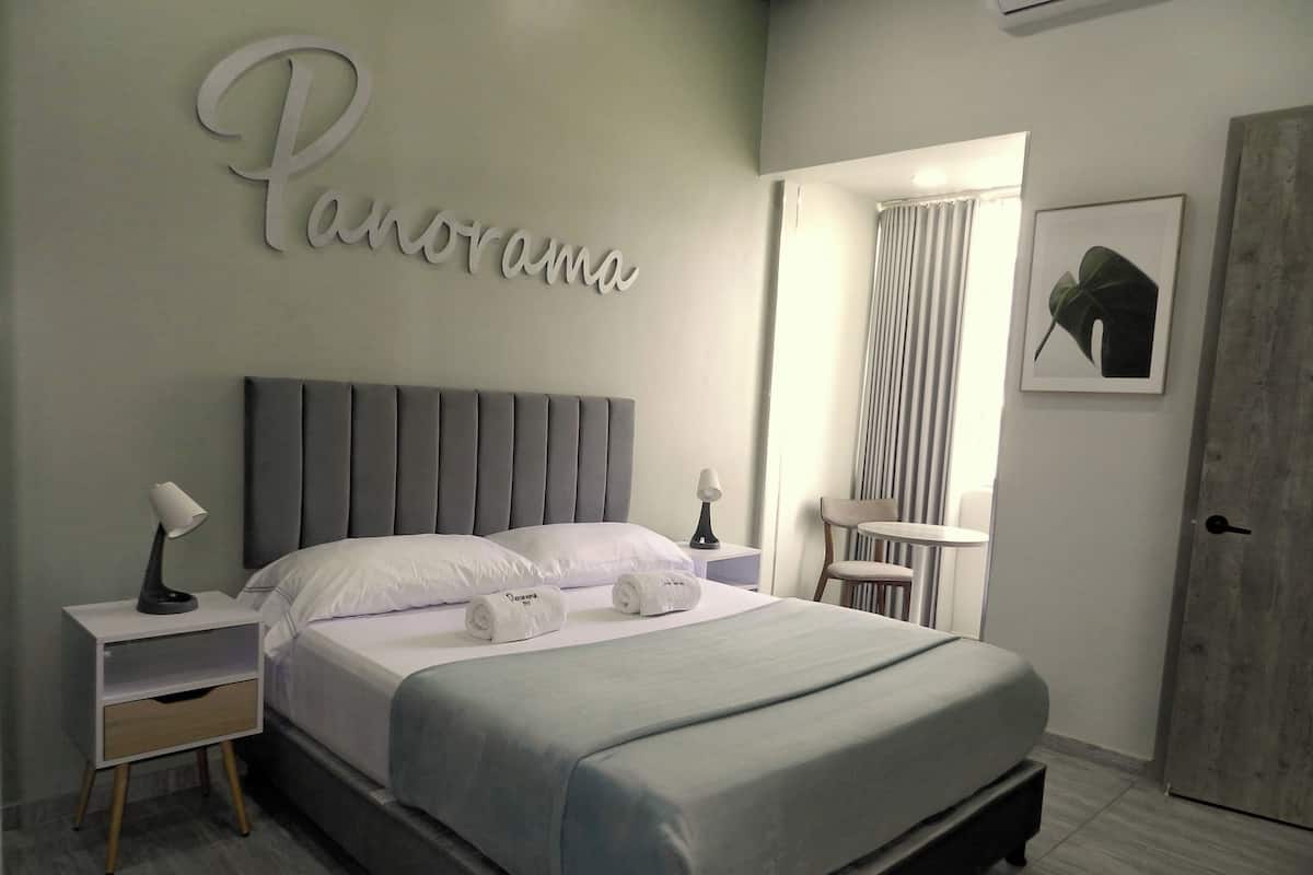 Un dormitorio moderno con cabecera gris, cama con ropa de cama blanca, dos mesitas de noche con lámparas, una mesita redonda pequeña y una puerta.
