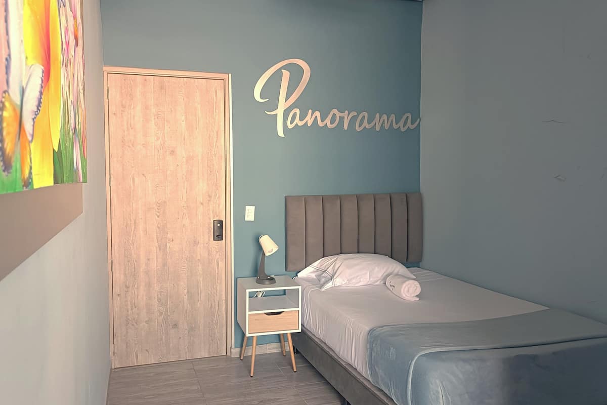Un dormitorio con cama, mesita de noche, lámpara, puerta de madera y pared con la palabra 'Panorama' escrita.