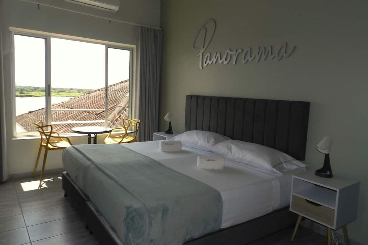 Habitación de hotel con cama, mesita de noche, silla, mesa y vista al río.