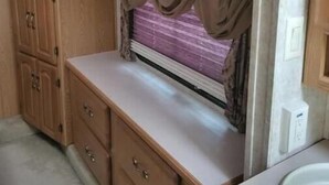 A compact kitchen with wooden cabinets, a countertop, and a window with curtains. - Spacious Clean RV Camper (Palm Bay)