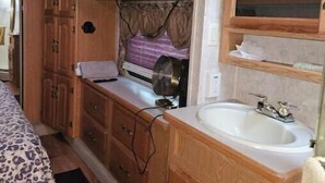 A compact RV interior with wooden cabinetry, a bed with a leopard print cover, a sink, and a television mounted above the cabinets. - Spacious Clean RV Camper (Palm Bay)