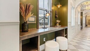 Un couloir avec un banc en bois, deux tabourets blancs rembourrés, une fenêtre avec une lampe et un vase décoratif contenant des plantes séchées.