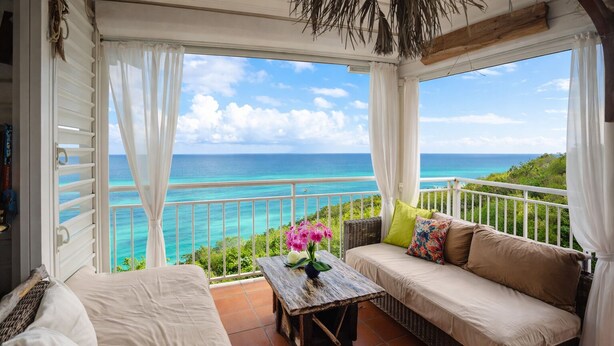 A balcony with a view of the ocean, a wicker sofa, a small table with a vase of flowers, and a white bed. - Anse des Rochers Beach & Pool Resort (Saint-François)