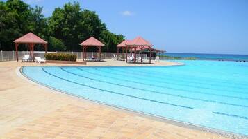 A swimming pool with lounge chairs and gazebos, surrounded by a tiled deck and a view of the ocean.