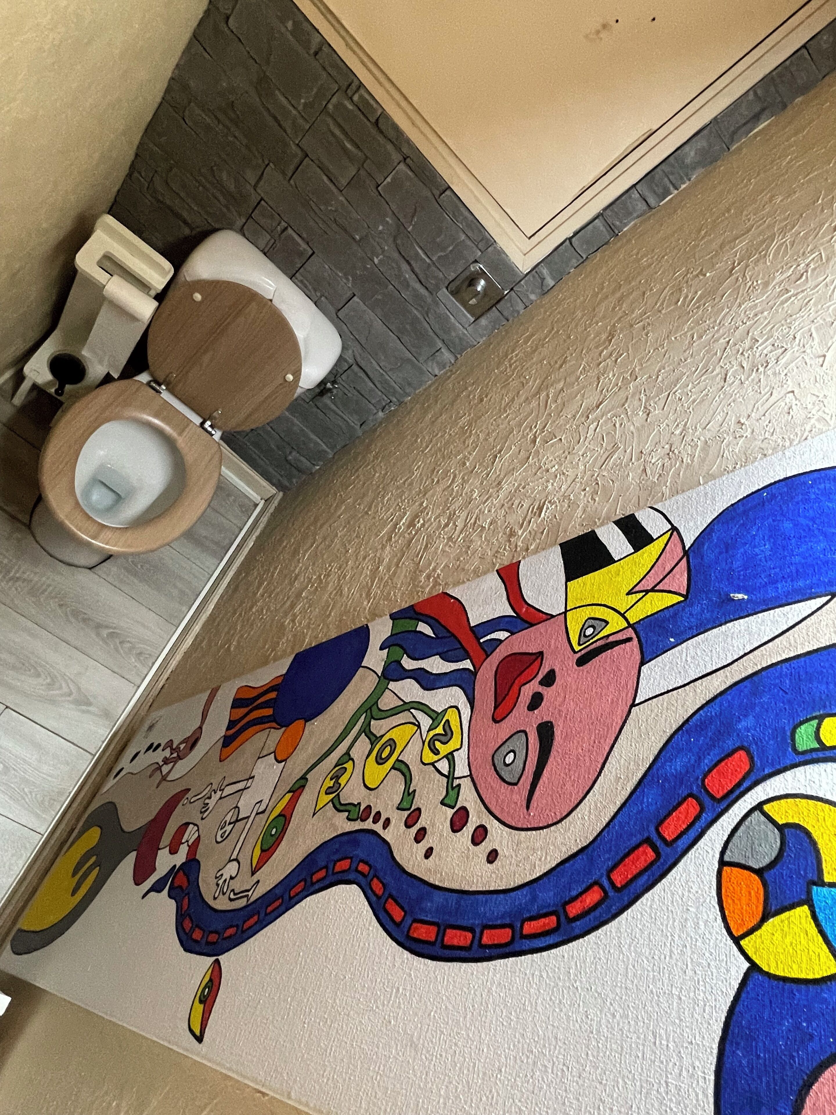 A bathroom with a wooden toilet, a tiled floor, and a colorful mural on the wall.