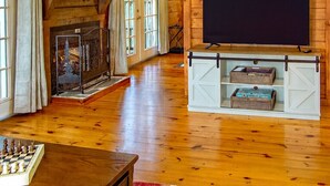 Een gezellig interieur van een chalet met een open haard, houten vloeren, een aan de muur bevestigde televisie en een schaakbord op een tafel.