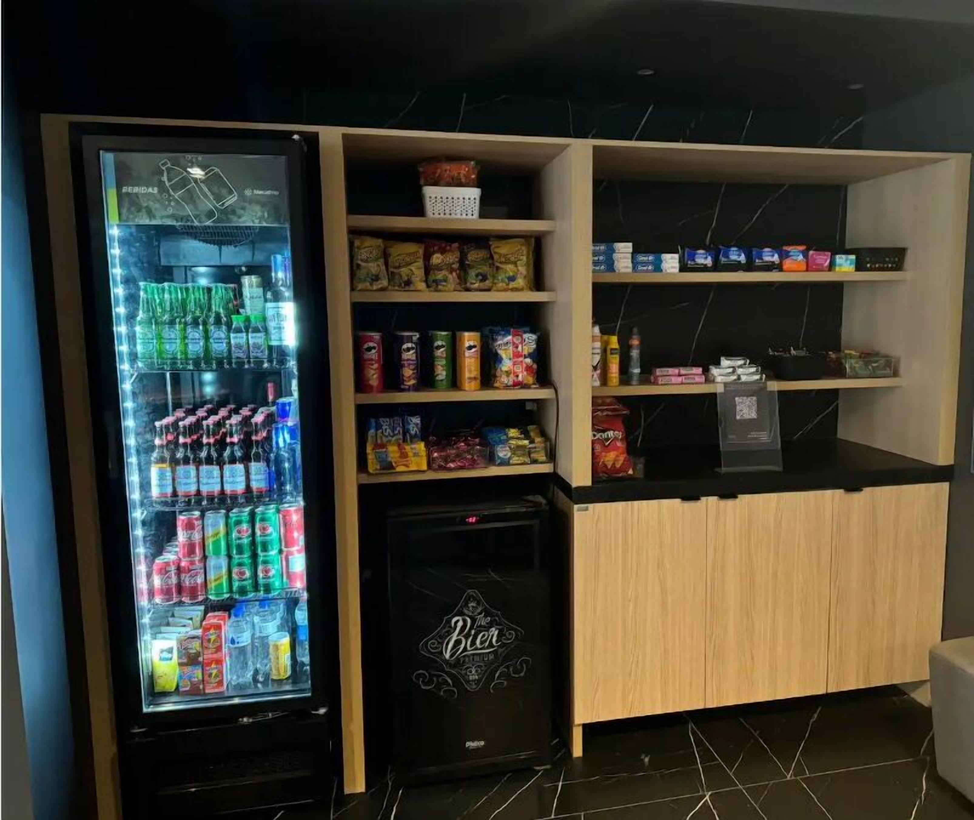 A vending machine stocked with various beverages and snacks, a refrigerator with a 'Bier Premium' logo, and shelves with packaged goods.