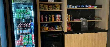 A vending machine stocked with various beverages and snacks, a refrigerator with a 'Bier Premium' logo, and shelves with packaged goods.