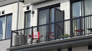 Un edificio de varios pisos con un balcón que tiene barandilla de metal y sillas rojas.