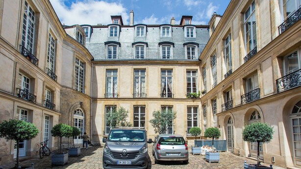Un patio con dos coches aparcados, árboles en macetas y un edificio con ventanas arqueadas y balcones.