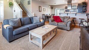 A modern living room with a grey sofa, a wooden coffee table, and a red throw blanket. A staircase leads to another level, and a kitchen area is visible in the background. - TWO townhomes for your giant group! (Corvallis)