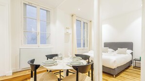 A room with a dining table set for four, a bed with a grey headboard, and two windows with curtains. - Cosy Apartment - 1br/4p - Near Champs-elysees (Paris)