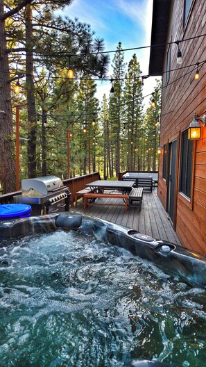 Una terraza de madera con un jacuzzi, asientos exteriores y luces colgantes.